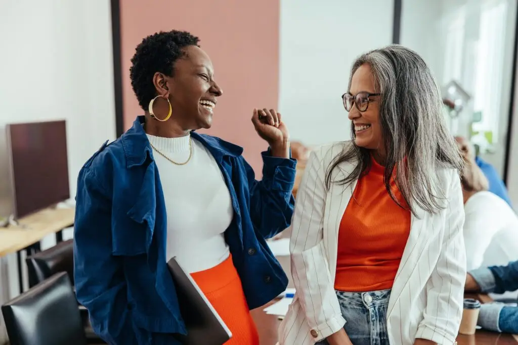 Two stylish women, one mature and one young, laughing together in a colorful and vibrant team workspace.